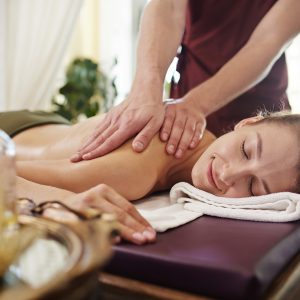 Portrait of beautiful young woman lying on massage table with eyes closed and smiling blissfully enjoying SPA treatment while man massaging her back with lotions and body oils.